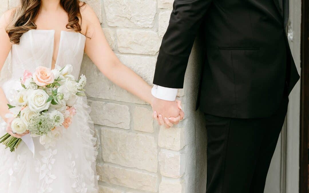 Bride and groom hold hands around a stone wall, both in formal attire.