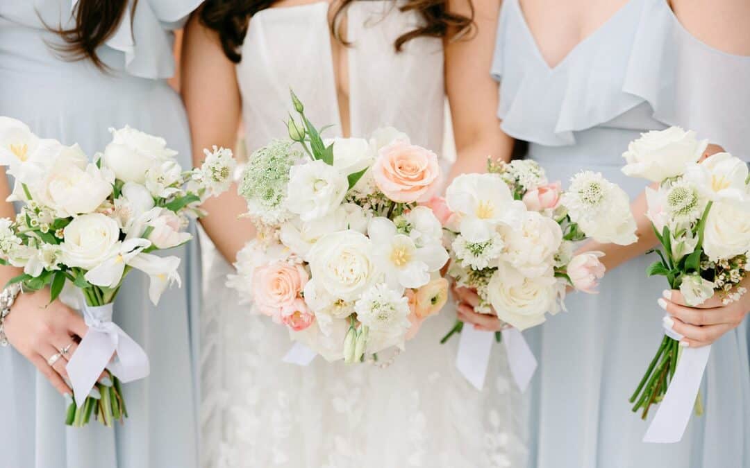 Three women in light blue dresses and a bride in white holding pastel bouquets featuring white and pink flowers.