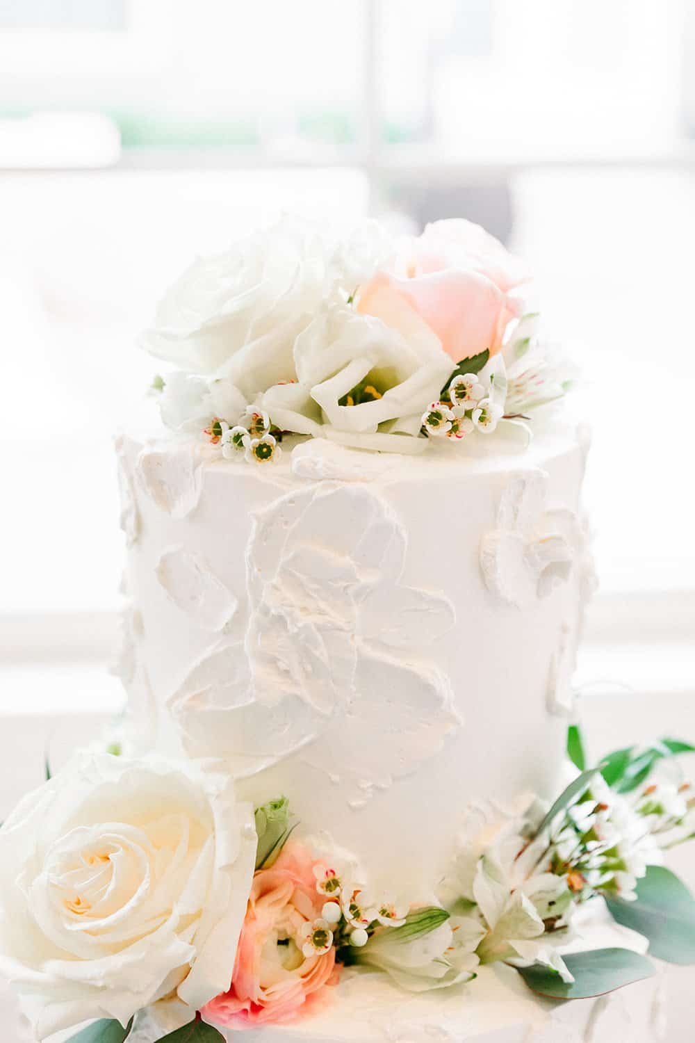 Two-tier white cake adorned with white and pink roses, with green leaves and small white flowers scattered around.