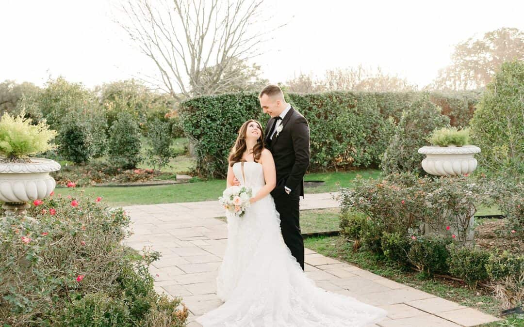 A couple in wedding attire stands on a stone path in a garden, with the bride holding a bouquet.