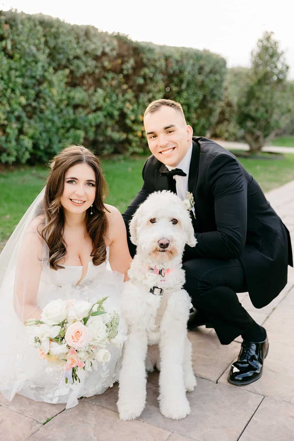 Bride and groom kneel beside a seated white dog in a wedding setting outdoors.