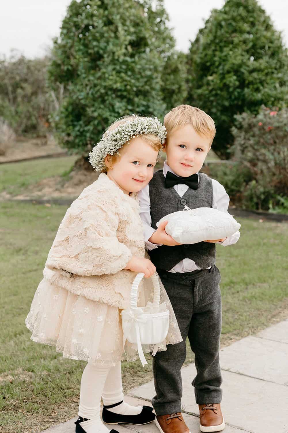 Two children dressed formally: a girl in a beige dress with a flower headband and a boy in a bow tie and vest holding a pillow, outdoors with greenery.