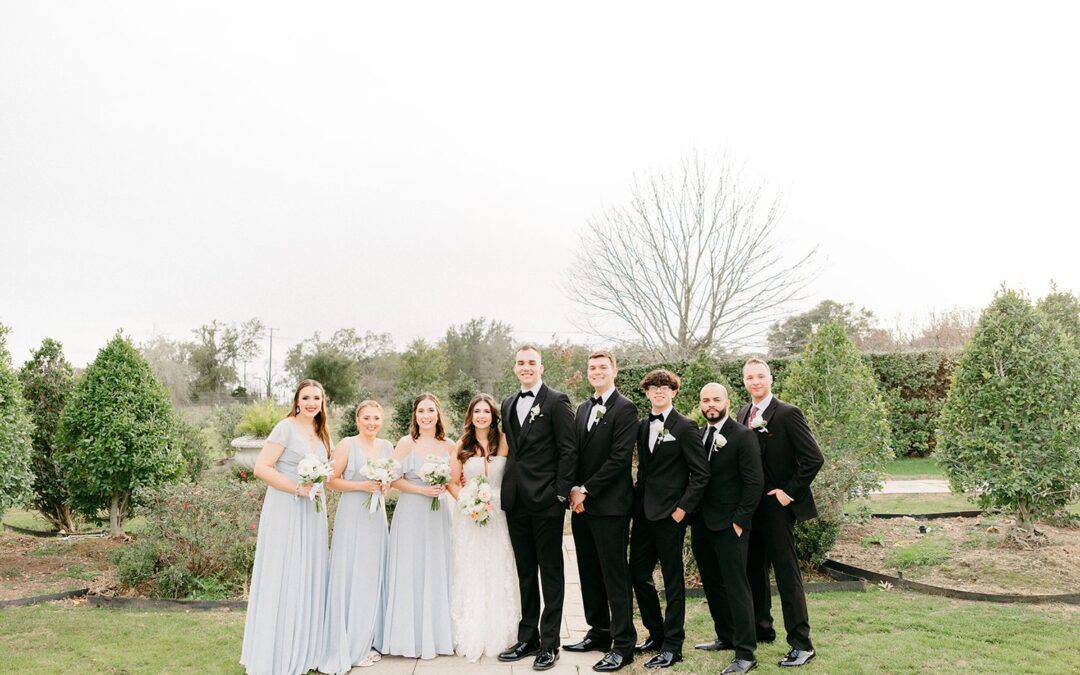 Wedding party outdoors with bridesmaids in light blue dresses, groomsmen in black suits, and the bride and groom in the center.