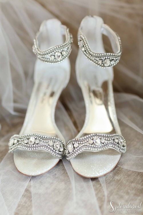 Close-up of elegant white high-heeled sandals adorned with intricate silver beadwork and rhinestones, set on a sheer fabric background.