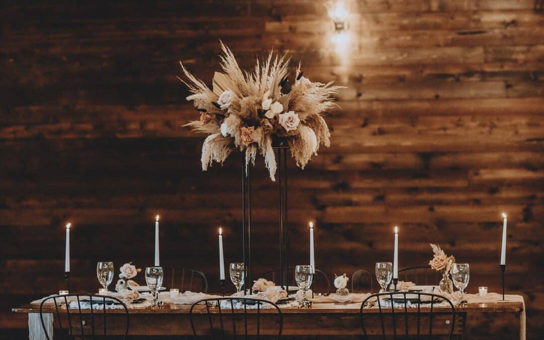 A rustic dining table is set for a formal event with tall candles, glassware, and a centerpiece of pampas grass and roses, against a wooden paneled background.