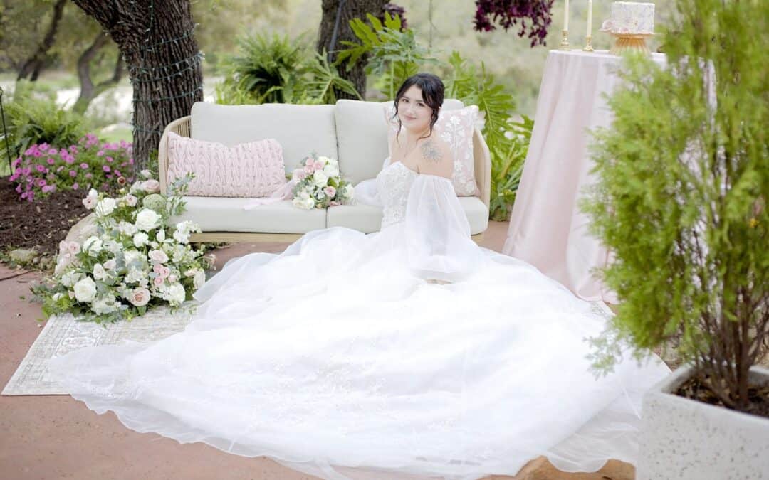 A bride in a white gown sits on a sofa outdoors, surrounded by flowers and greenery. A table with a cake is beside her.