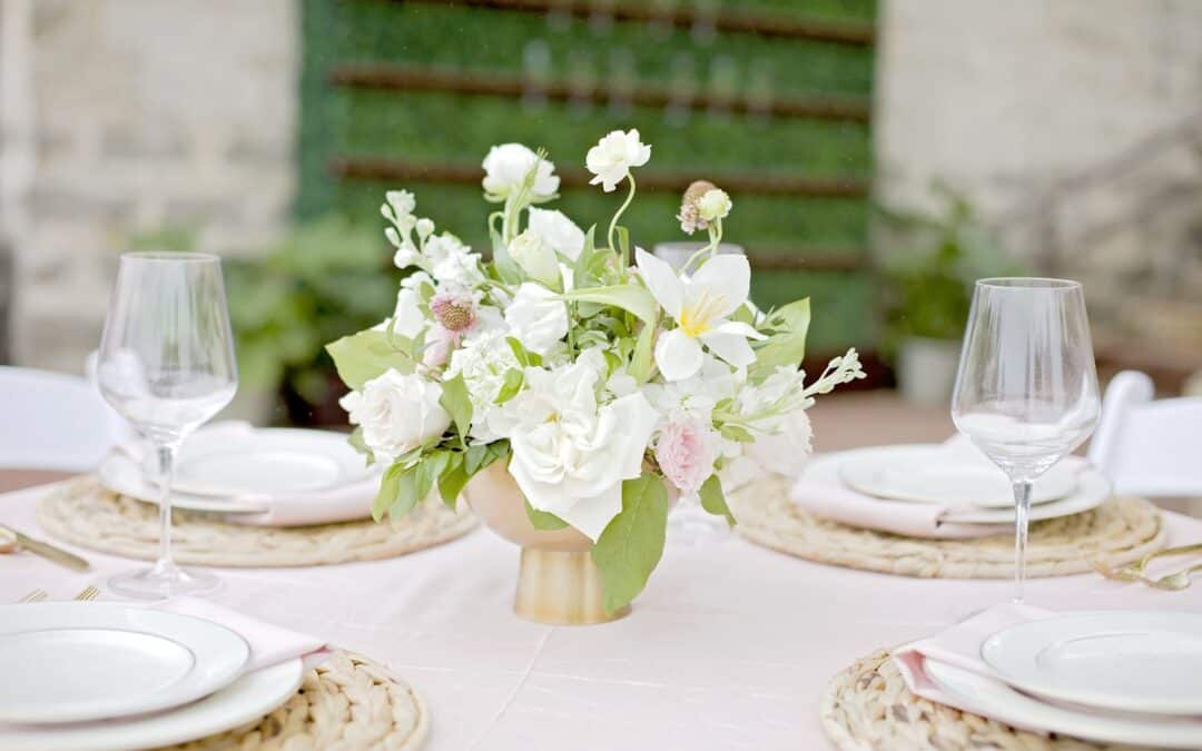 A table is set outdoors with a floral centerpiece, two empty wine glasses, and plates on woven placemats.