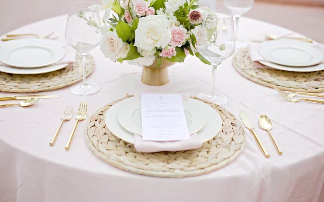 Table with woven placemats, white plates, gold utensils, a tall floral centerpiece, wine glasses, and a menu on one plate.